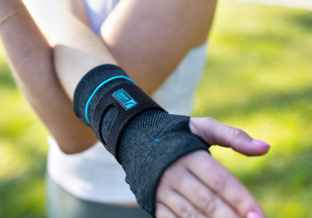 En person bär en svart handledsskydd märkt "Formfit" i blått, medan de står utomhus på en grönskande plats. Handlederna är placerade i en bekväm position, med solljus som lyser mjukt över scenen.